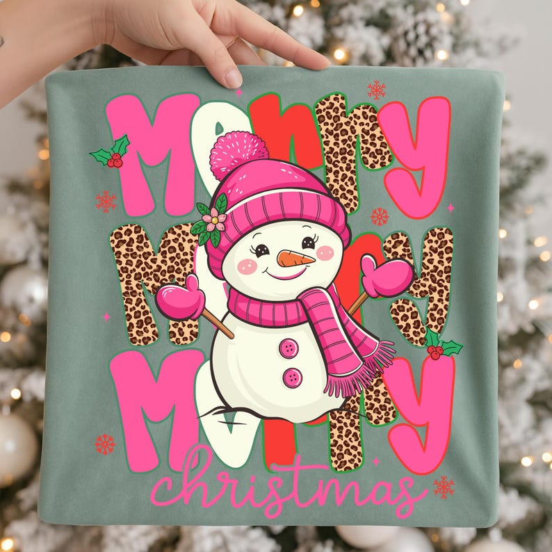 May include: A green pillow with a festive design featuring a snowman wearing a pink hat, scarf, and mittens. The words "Merry Christmas" are displayed in pink and leopard print, with red accents and snowflakes.