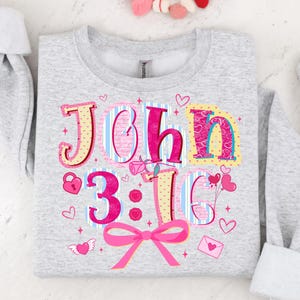 May include: Pale grey sweatshirt featuring the text "John 3:16" in a colourful, decorative font. The design incorporates hearts, a pink bow, and a rose. The sweatshirt is a comfortable, casual style.