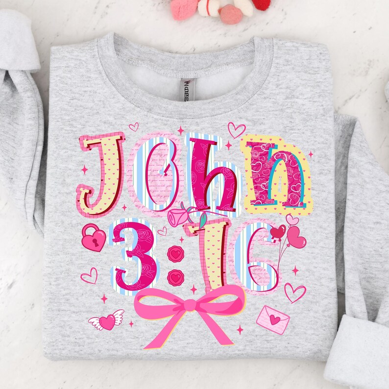 May include: Pale grey sweatshirt featuring the text "John 3:16" in a colourful, decorative font. The design incorporates hearts, a pink bow, and a rose. The sweatshirt is a comfortable, casual style.