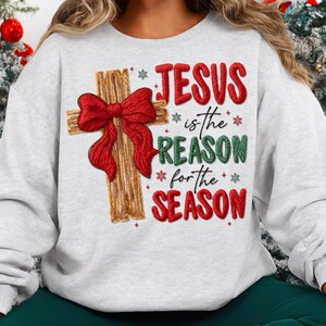 May include: A light gray sweatshirt with a Christmas theme. The design features a wooden cross adorned with a red bow. Text on the sweatshirt reads "JESUS is the REASON for the SEASON" in red and green.