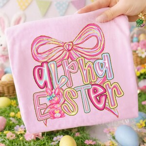 May include: A pink t-shirt featuring the text "Alpha Easter" in a colorful, outlined font, along with a bow and a bunny graphic. The design has a pastel color scheme with gold glitter accents, perfect for Easter.