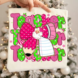 Peut inclure: T-shirt cr&egrave;me avec un motif de No&euml;l. Le graphisme comprend le mot "Teacher" en rose et vert, un n&oelig;ud rose &agrave; pois, un crayon et une illustration du P&egrave;re No&euml;l. Des flocons de neige et des accents de houx compl&egrave;tent le th&egrave;me festif.