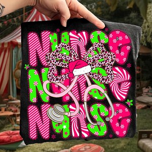 May include: Black t-shirt with the word "NURSE" in pink and green, decorated with a bow, Santa hat, and stethoscope. The bow has a leopard print design. The letters are decorated with stripes, stars, and snowflakes.