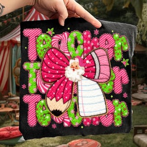 May include: Black t-shirt with a festive design featuring the words "Teacher Team" in pink and green candy cane-style lettering. A large pink bow with a Santa Claus figure and a pencil is in the center. The design includes snowflakes and stars.