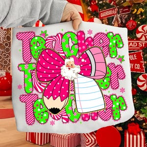 Peut inclure: Un sac fourre-tout blanc avec le mot "Teacher" en rose et vert, avec un motif de crayon du P&egrave;re No&euml;l. Le motif comprend un n&oelig;ud rose &agrave; pois blancs, un crayon et des flocons de neige. Le sac est tenu devant un sapin de No&euml;l.