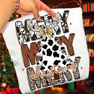 May include: White Christmas-themed t-shirt with the words "MERRY CHRISTMAS" in a mix of fonts and patterns. The design includes a cow-print Christmas tree, antlers, and a gold bow. The text is in brown, black, and leopard print.