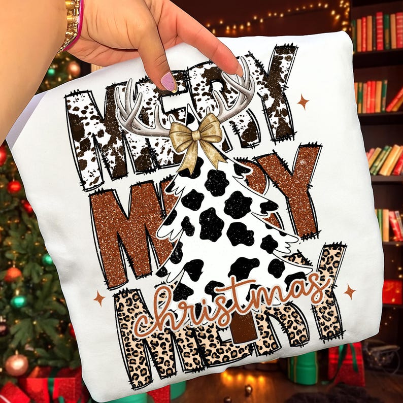 May include: White Christmas-themed t-shirt with the words "MERRY CHRISTMAS" in a mix of fonts and patterns. The design includes a cow-print Christmas tree, antlers, and a gold bow. The text is in brown, black, and leopard print.