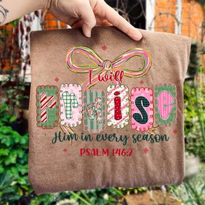 May include: A brown t-shirt with the words "I will Praise Him in every season" in a decorative font. The word "Praise" is spelled out in colorful, patterned letters. The text "PSALM 146:2" is also printed on the shirt.