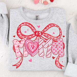 May include: A light gray sweatshirt with a large red bow and the word "MOM" in a decorative font. The letters are adorned with hearts and patterns in shades of pink and red, perfect for Valentine's Day or Mother's Day.