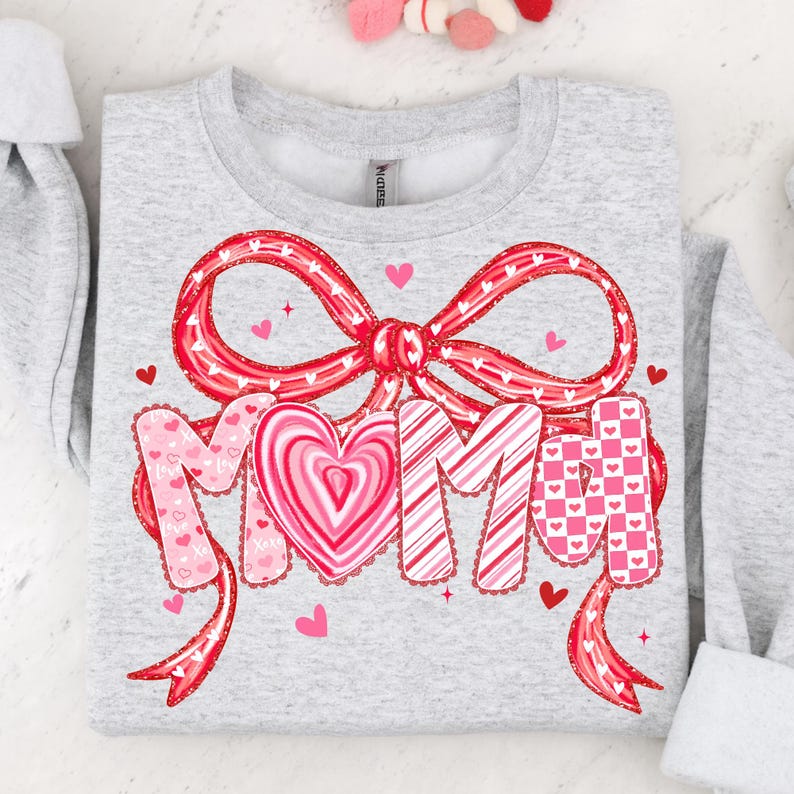 May include: A light gray sweatshirt with a large red bow and the word "MOM" in a decorative font. The letters are adorned with hearts and patterns in shades of pink and red, perfect for Valentine's Day or Mother's Day.