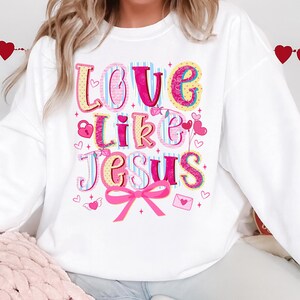 May include: White sweatshirt with the phrase "Love Like Jesus" in colorful, whimsical lettering. The design includes hearts, a bow, and other decorative elements, perfect for a casual, faith-based style.