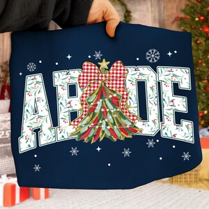 May include: Navy blue pillow with the word "ABODE" in white, decorated with a Christmas tree design. The tree is adorned with red and green details, a red and white checkered bow, and a gold star. Snowflakes and stars are scattered around the text.
