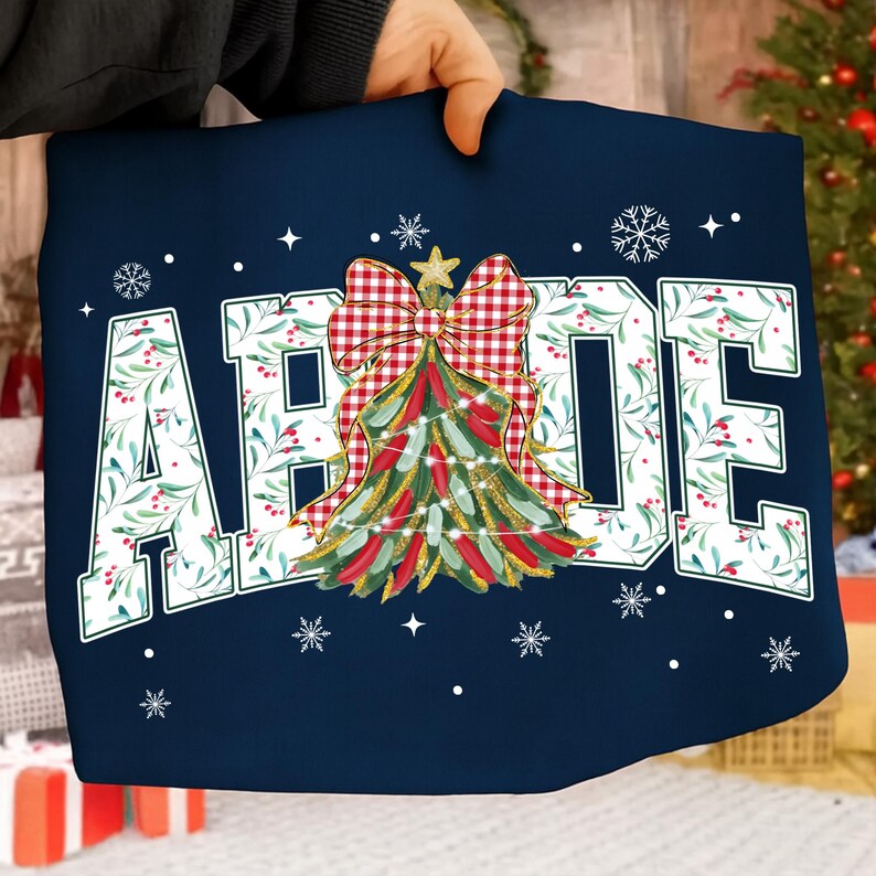 May include: Navy blue pillow with the word "ABODE" in white, decorated with a Christmas tree design. The tree is adorned with red and green details, a red and white checkered bow, and a gold star. Snowflakes and stars are scattered around the text.