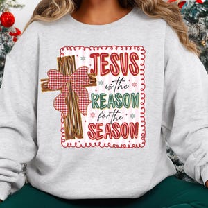 May include: A light gray sweatshirt featuring a decorative design with the text "JESUS is the REASON for the SEASON." The design includes a wooden cross with a red and white plaid bow. The text is in red, green, and white.