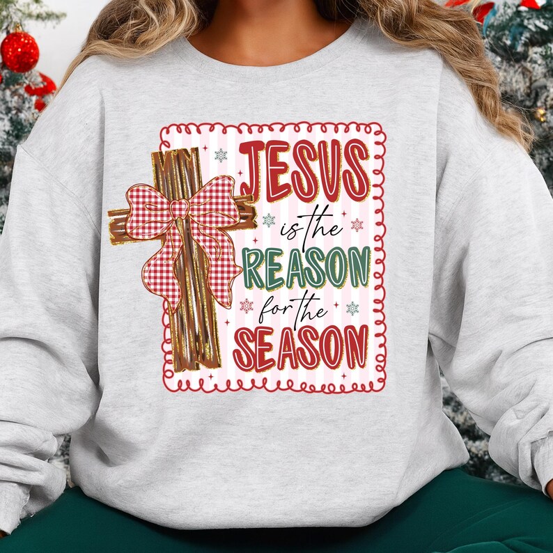 May include: A light gray sweatshirt featuring a decorative design with the text "JESUS is the REASON for the SEASON." The design includes a wooden cross with a red and white plaid bow. The text is in red, green, and white.