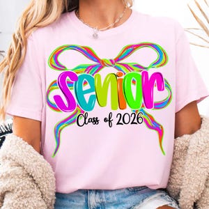 May include: A pale pink t-shirt with a colourful bow design and the word "Senior" in bright, multi-coloured letters. Below the word "Senior" is the text "Class of 2026" in black. The shirt is a casual style.