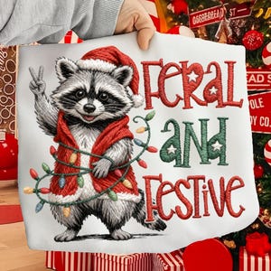 May include: White fabric with a raccoon wearing a Santa hat and red vest, holding up a peace sign. The text "FERAL AND FESTIVE" is embroidered in red and green with star accents. A string of colorful lights is wrapped around the raccoon.