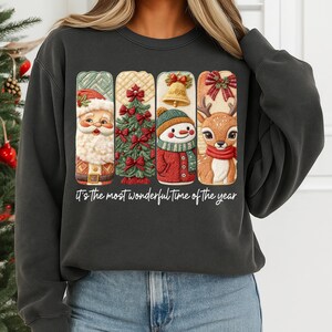 May include: Dark gray sweatshirt with a festive Christmas design. The graphic features Santa Claus, a Christmas tree, a snowman, and a reindeer, each in a rectangular frame. The text "It's the most wonderful time of the year" is printed below the design.