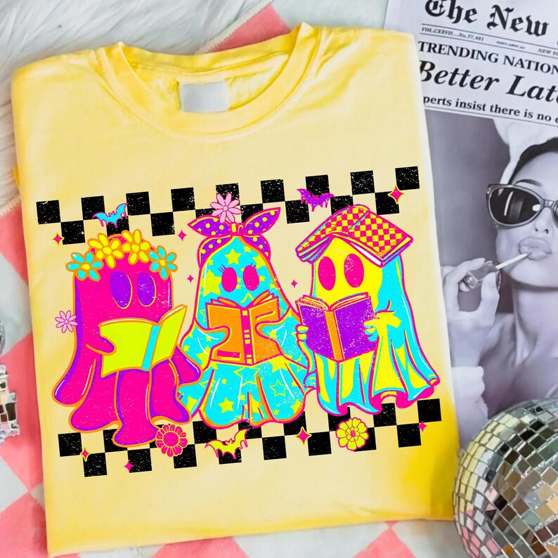 May include: Yellow t-shirt featuring a colorful graphic of three ghost-like figures reading books. The figures are outlined in black and have vibrant pink, blue, and yellow details. A black and white checkered pattern borders the top of the design.