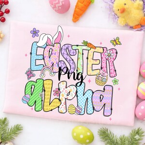 Op de afbeelding: Een lichtroze t-shirt met de woorden "Easter Alpha Png" in kleurrijke, patroonletters. Het ontwerp bevat paaseieren, een regenboog, vlinders en een wortel. Paas-thema kleding.