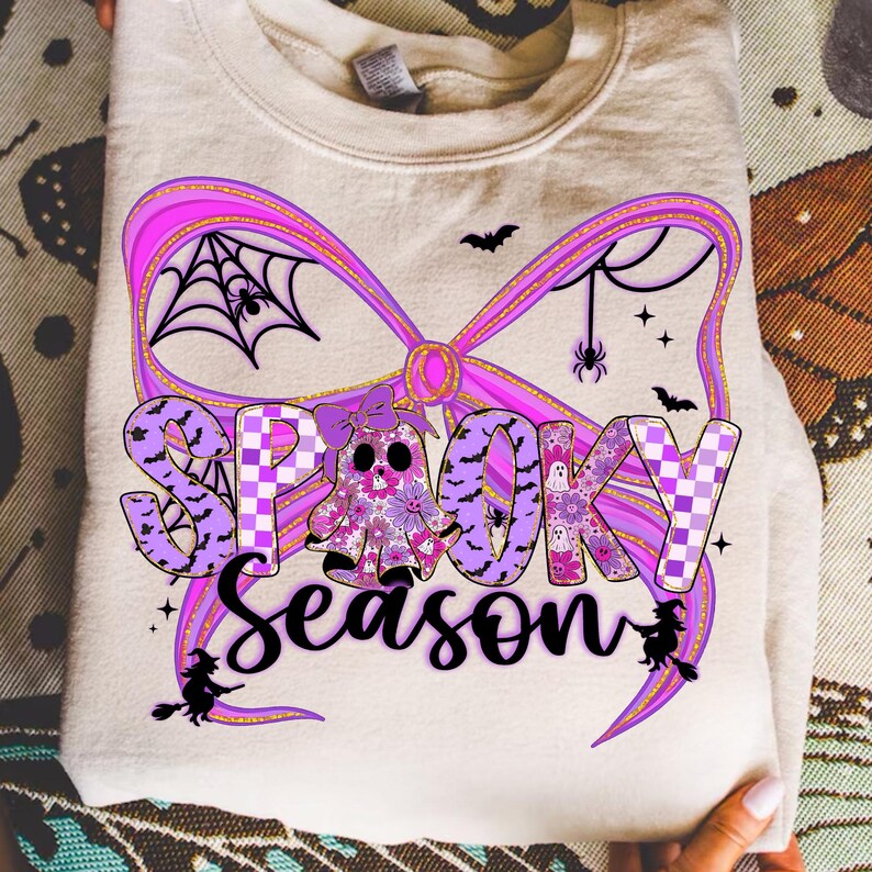 May include: A folded cream sweatshirt with a purple bow design and the words "Spooky Season". The design includes bats, spiders, and a ghost with floral details. A seasonal item for Halloween.