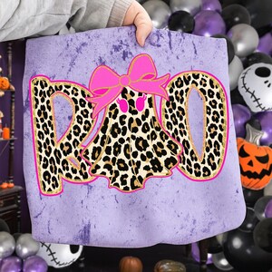 May include: A lavender tote bag with the word "RAD" in leopard print letters outlined in gold. A ghost in a leopard print costume with a pink bow is in the center. The bag has a distressed look, perfect for Halloween.
