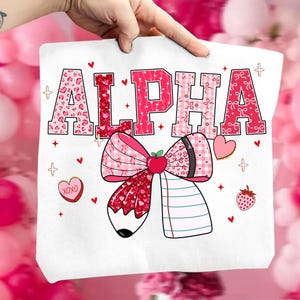 Op de afbeelding: Witte draagtas met het woord "ALPHA" in roze en rood gekleurde letters. Onder de tekst staat een grote strik met een potloodontwerp, samen met harten, aardbeien en de tekst "XOXO".