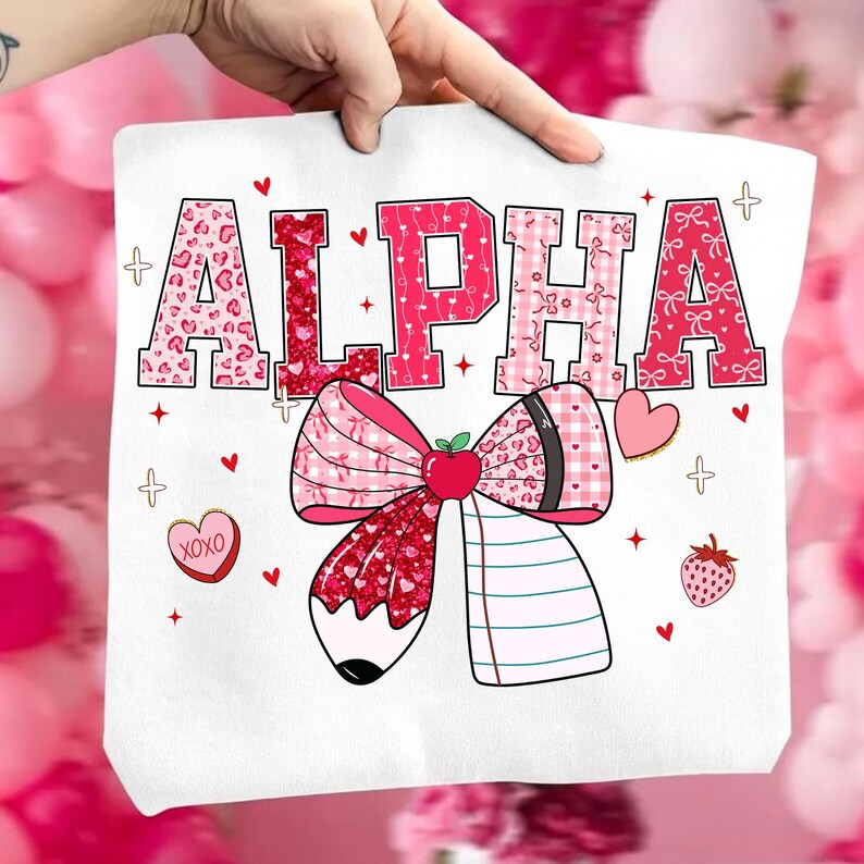 Op de afbeelding: Witte draagtas met het woord "ALPHA" in roze en rood gekleurde letters. Onder de tekst staat een grote strik met een potloodontwerp, samen met harten, aardbeien en de tekst "XOXO".