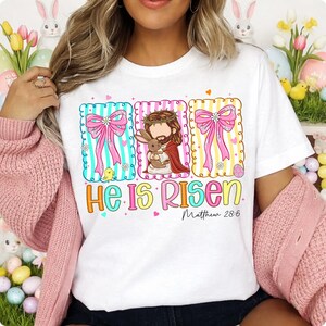 May include: White t-shirt featuring an Easter-themed design. The graphic includes pink bows, a depiction of Jesus with a rabbit, and the text "He Is Risen" with the Bible verse Matthew 28:6.