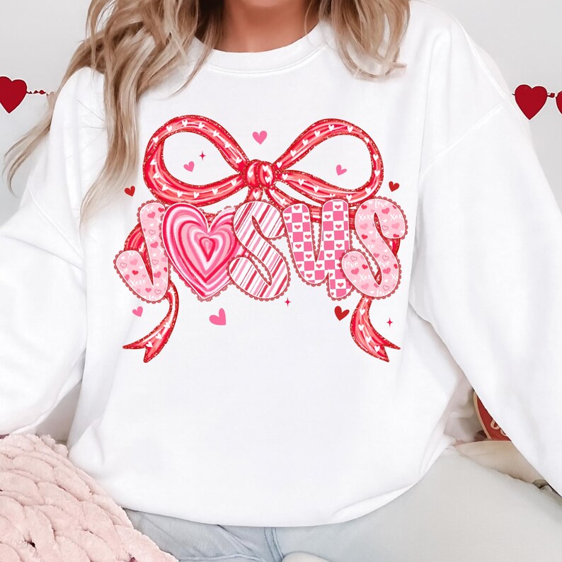 May include: White sweatshirt with a large graphic featuring the word "JESUS" in pink letters, a pink heart, and a red bow. The design includes hearts and patterns. The sweatshirt is a casual style.