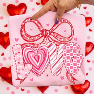 May include: A pale pink t-shirt featuring a large red bow and the word "LOVE" in a decorative font. The "O" is a heart shape. The shirt has small heart and "XOXO" details, ideal for Valentine's Day.
