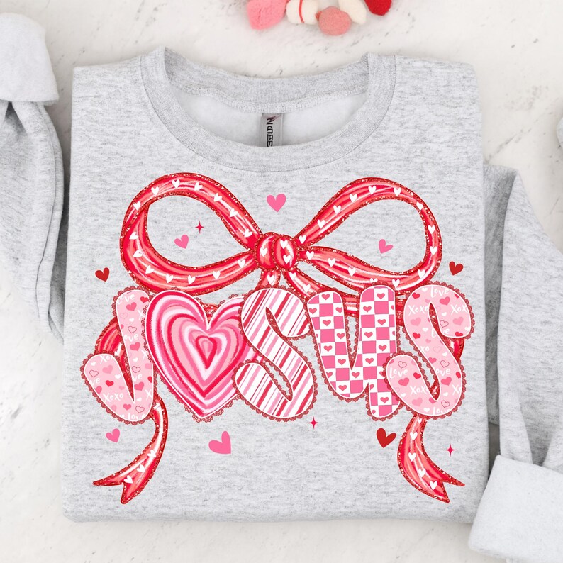 May include: A light gray sweatshirt with a large red bow and the word "JESUS" in pink and white lettering. The letters are decorated with hearts and the sweatshirt is a Valentine's Day theme.