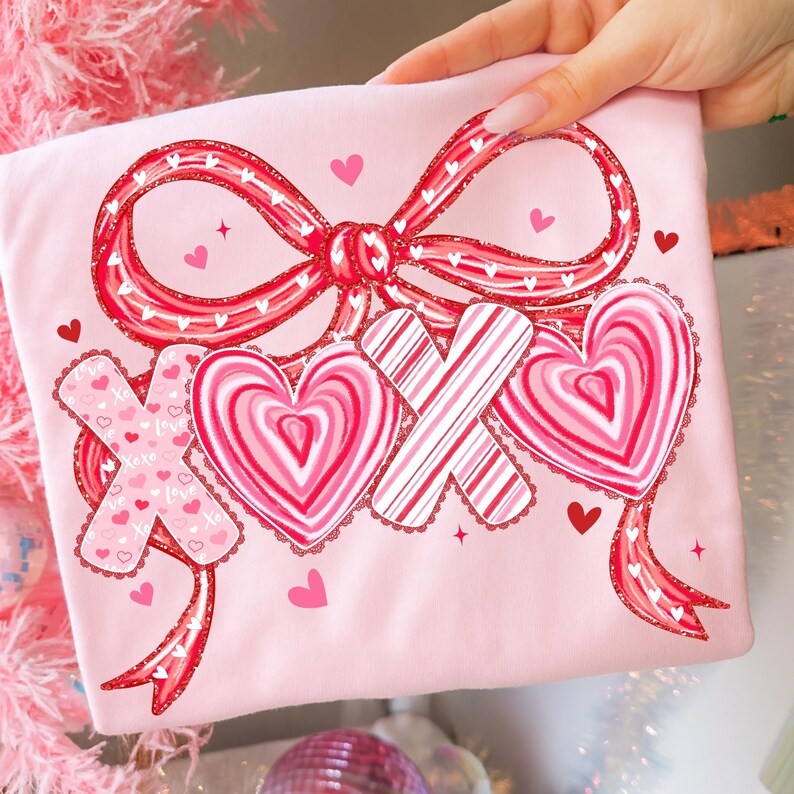 May include: A pale pink t-shirt with a large red bow and glitter detailing. The design includes two hearts and two X's in pink and red, with a lace trim. Small hearts are scattered around the design, creating a romantic theme.