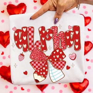 May include: White t-shirt with the word "ALPHA" in red, heart-filled letters. A large red bow with a checkered pattern and hearts is below the text, along with a pencil and strawberry graphic. Small hearts and crosses are scattered around the design.