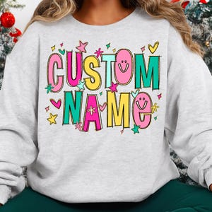 May include: A light gray sweatshirt with the words "CUSTOM NAME" in a colorful, playful font. The text is outlined in black and decorated with stars, hearts, and smiley faces. The sweatshirt is a casual, comfortable style.