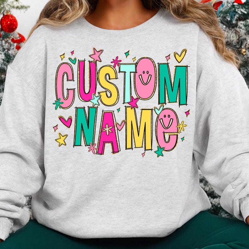 May include: A light gray sweatshirt with the words "CUSTOM NAME" in a colorful, playful font. The text is outlined in black and decorated with stars, hearts, and smiley faces. The sweatshirt is a casual, comfortable style.