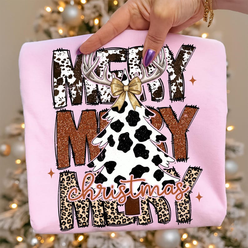May include: A light pink sweatshirt with a festive Christmas design. The design features the words "MERRY CHRISTMAS" in a mix of fonts and textures, including cow print, glitter, and leopard print. A Christmas tree with a cow print pattern is in the center.