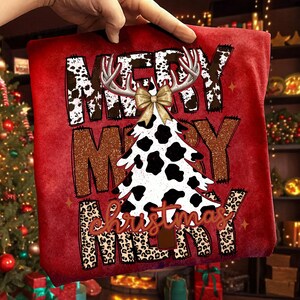 May include: Red Christmas-themed t-shirt with the words "MERRY CHRISTMAS" in a mix of fonts and patterns. The design includes a cow-print Christmas tree, antlers, and a bow.
