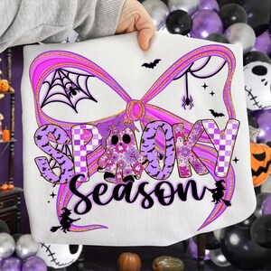 May include: White sweatshirt featuring a purple bow design and the words "Spooky Season". The design includes a ghost, bats, spiders, and a spiderweb, ideal for Halloween celebrations.