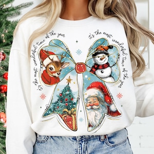 May include: White long-sleeve sweatshirt featuring a large bow design with Christmas-themed illustrations. The bow includes a reindeer, snowman, Christmas tree, and Santa Claus. Text around the bow reads "It's the most wonderful time of the year."
