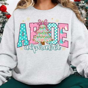 May include: A light gray sweatshirt featuring a colorful graphic design. The design spells out "AFDE" with each letter in a different pattern, including stars and a checkered pattern. A Christmas tree is in the center, and the word "alphabet" is written below.