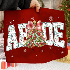 May include: A red Christmas-themed design featuring the word "ABODE" in white letters with a green floral pattern. A Christmas tree with a red and white checkered bow is in the center, with snowflakes and stars scattered around.