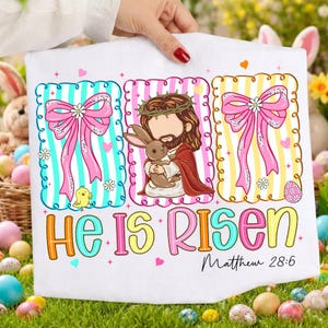 May include: White Easter t-shirt with three framed designs. The shirt features a Jesus illustration holding a bunny, and two frames with bows and stripes. The text "He Is Risen" and "Matthew 28:6" are also displayed.