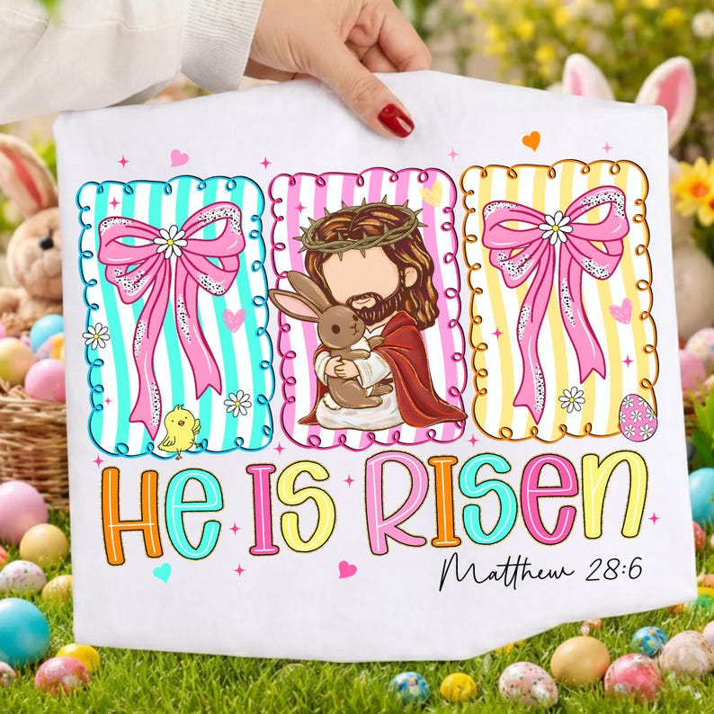 May include: White Easter t-shirt with three framed designs. The shirt features a Jesus illustration holding a bunny, and two frames with bows and stripes. The text "He Is Risen" and "Matthew 28:6" are also displayed.