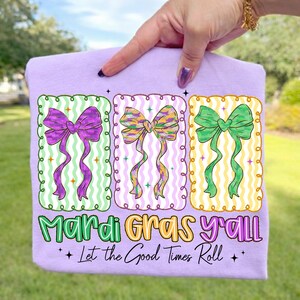 May include: A light purple t-shirt featuring three colorful bow designs in green, purple, and yellow. The text "Mardi Gras y'all" and "Let the Good Times Roll" are printed below the bows. The shirt is held up by a hand.