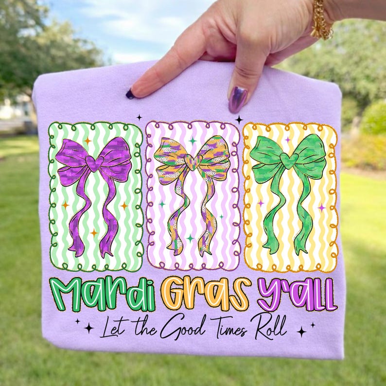 May include: A light purple t-shirt featuring three colorful bow designs in green, purple, and yellow. The text "Mardi Gras y'all" and "Let the Good Times Roll" are printed below the bows. The shirt is held up by a hand.