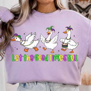 May include: A light purple sweatshirt featuring a graphic of three white geese in Mardi Gras attire. The geese are wearing jester hats and necklaces, with one playing a drum. The text "Let the Good Times Roll" is below.