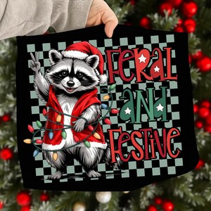 May include: Black square with a raccoon wearing a Santa hat and red vest, holding up a peace sign. The text "Feral and Festive" is in red and green, with a string of Christmas lights.
