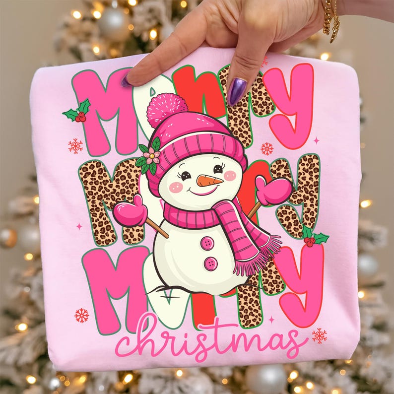 May include: Pink sweatshirt with a snowman graphic and the words "Merry Christmas". The snowman wears a pink hat, scarf, and mittens. The letters "M" and "Y" are in pink, and the letters "R" and "M" are in red. Some letters have a leopard print.
