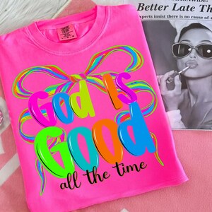 May include: Bright pink t-shirt with the phrase "God is Good" in colorful bubble letters and a rainbow bow design. The words "all the time" are printed below the main text. A Comfort Colors tag is visible.
